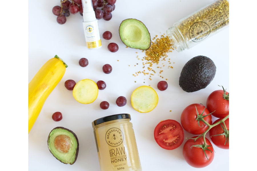 Jar of Beekeeper's Naturals raw honey with tomatoes, avocado, yellow squash, grapes, and spilled bee pollen on white