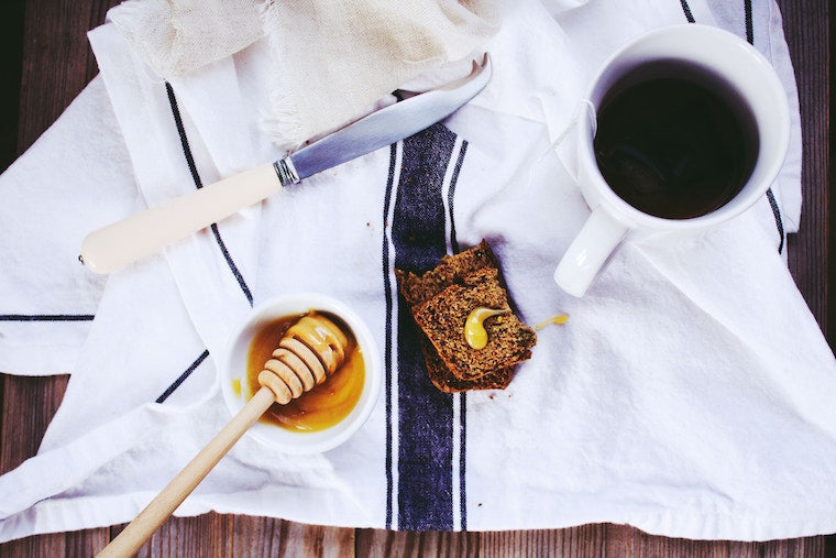 Honey dipper in a bowl of honey beside a knife, toast with honey drizzle and a coffee mug on a white striped towel