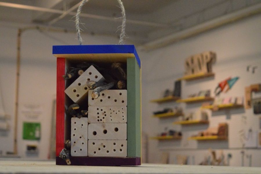 Front view of a small colorful wooden bee hotel showing drilled nesting blocks and bundled reeds, hanging in a craft workshop with blurred shelves in the background
