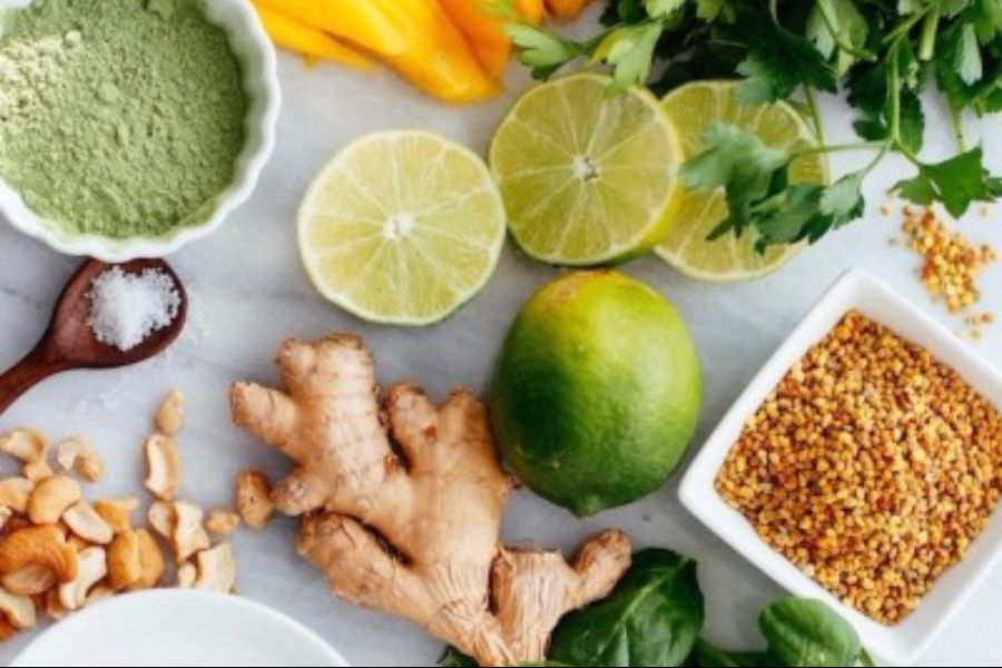Smoothie ingredients on a light surface: ginger, halved limes, bee pollen, cashews, matcha powder and leafy greens
