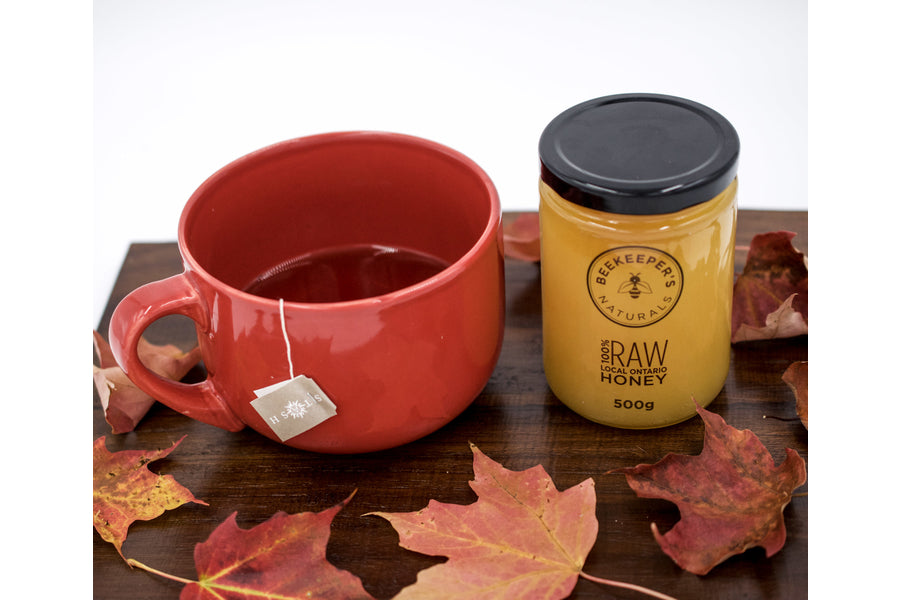 Front view of Beekeeper's Naturals 500 g raw local honey jar beside a red mug of tea on a wooden board with fall leaves