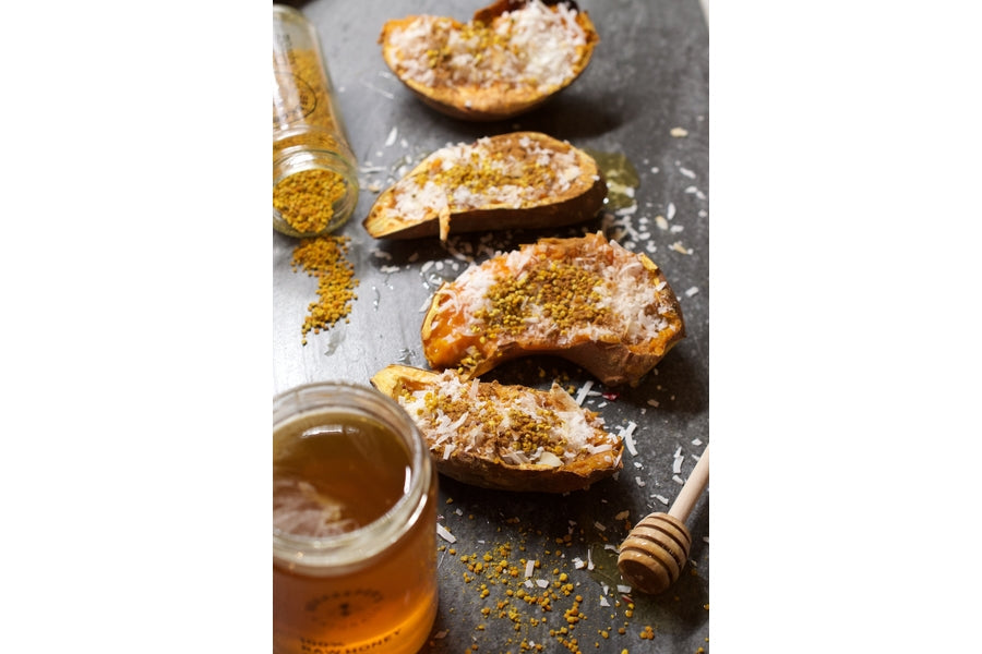 Roasted sweet potato halves with shredded coconut and bee pollen next to a jar of honey and dipper on dark slate