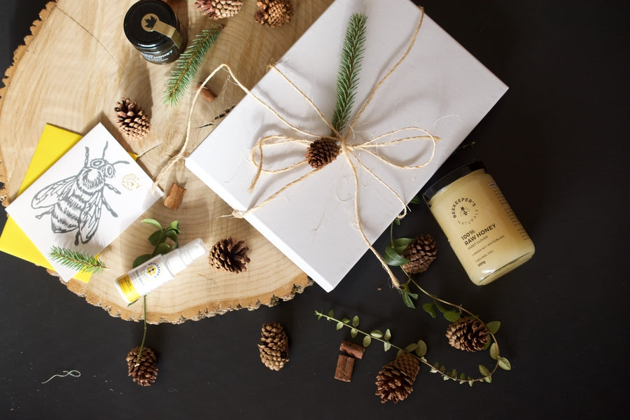 Flat lay of gift items with jar of raw honey, white wrapped box, roll-on balm, greeting card and pinecones