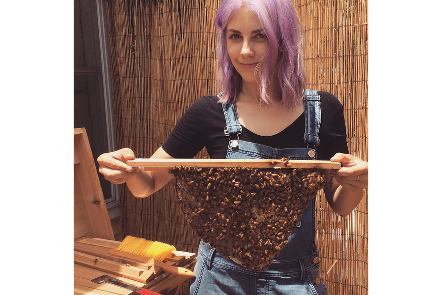 Woman with purple hair wearing denim overalls holding a honeycomb frame covered in bees in front of a bamboo screen
