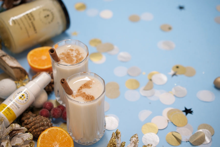 Two creamy cocktails with cinnamon sticks on a blue surface, surrounded by orange slices, pinecones, confetti, and a Beekeeper's Naturals bottle