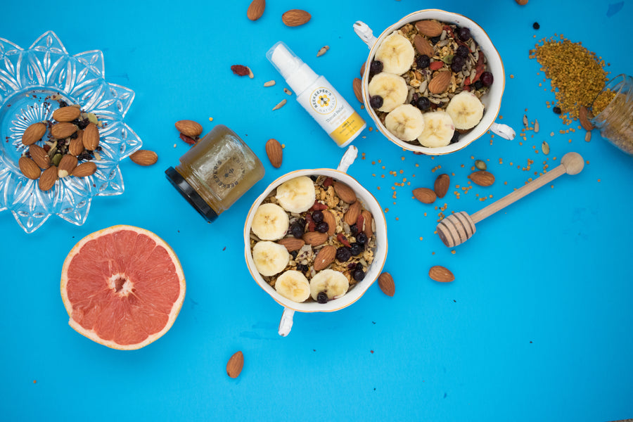 Two bowls of granola with banana slices, almonds and dried berries on a bright blue surface with a jar of honey, honey dipper and half a grapefruit