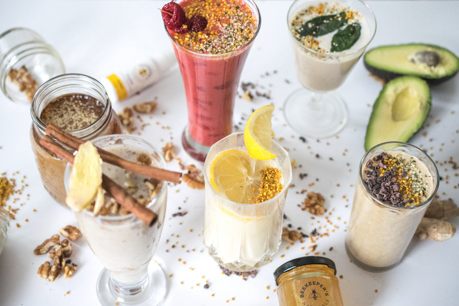 Assorted smoothies in glasses on a white table with bee pollen and garnishes, alongside avocado halves and a jar of honey