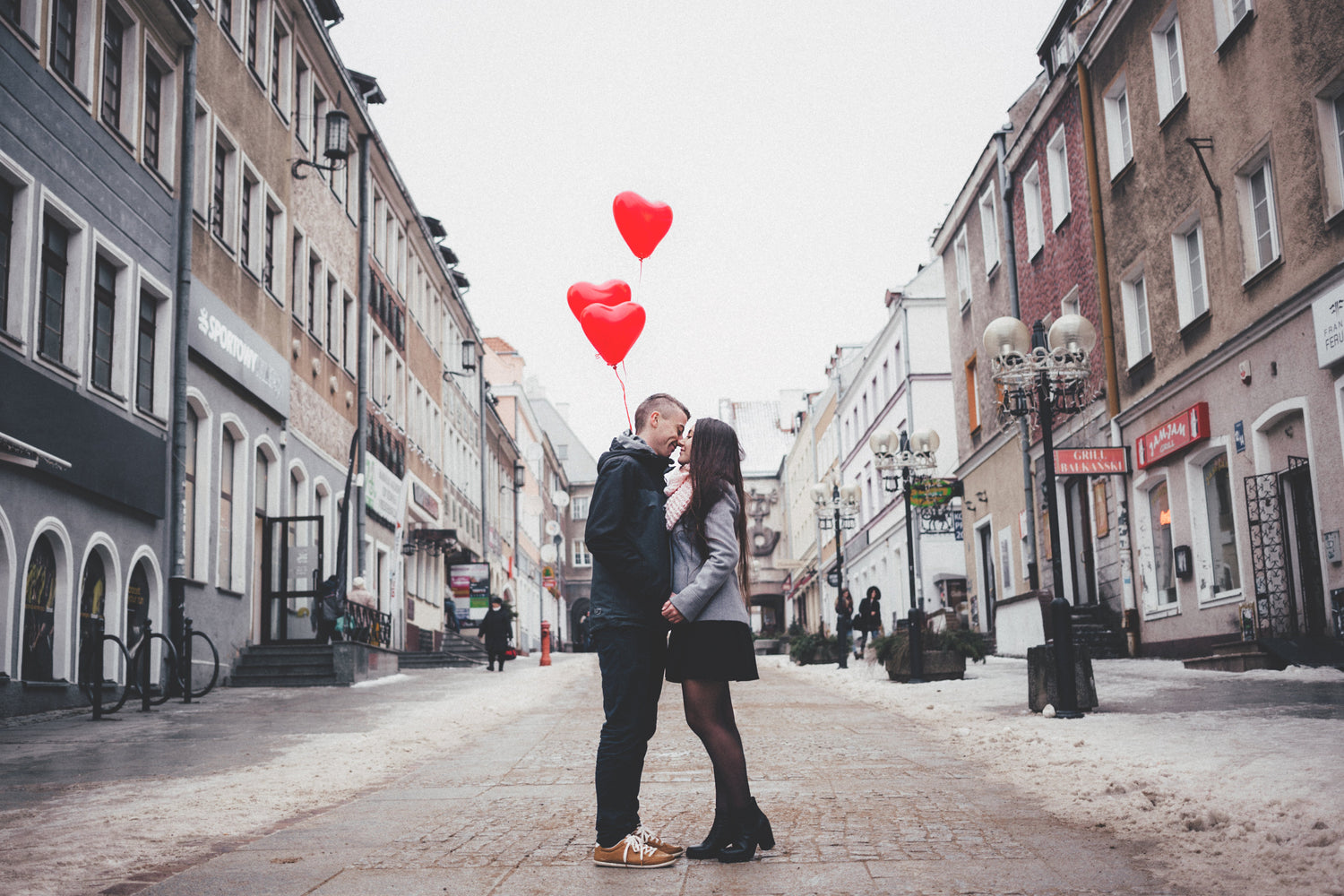 Couple embracing and about to kiss on a snowy city street with three red heart-shaped balloons overhead