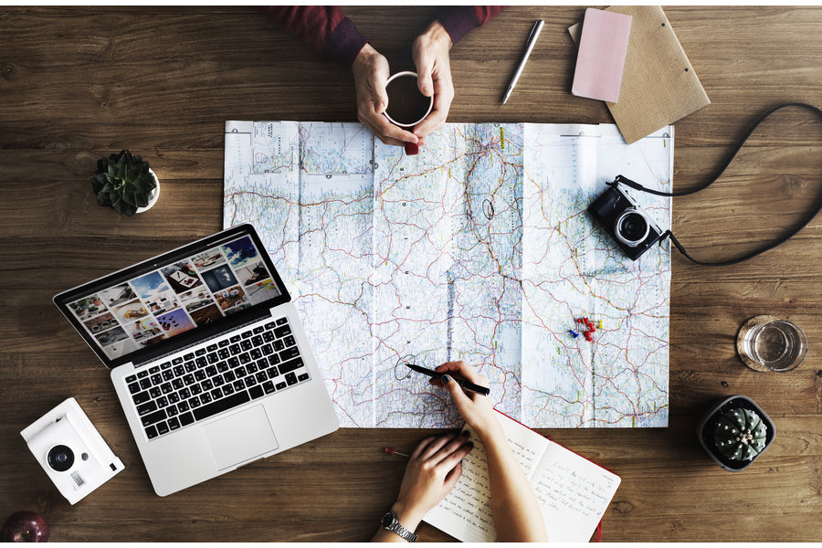 Hands marking a paper map while a laptop, camera, notebook, and coffee sit on a wooden table