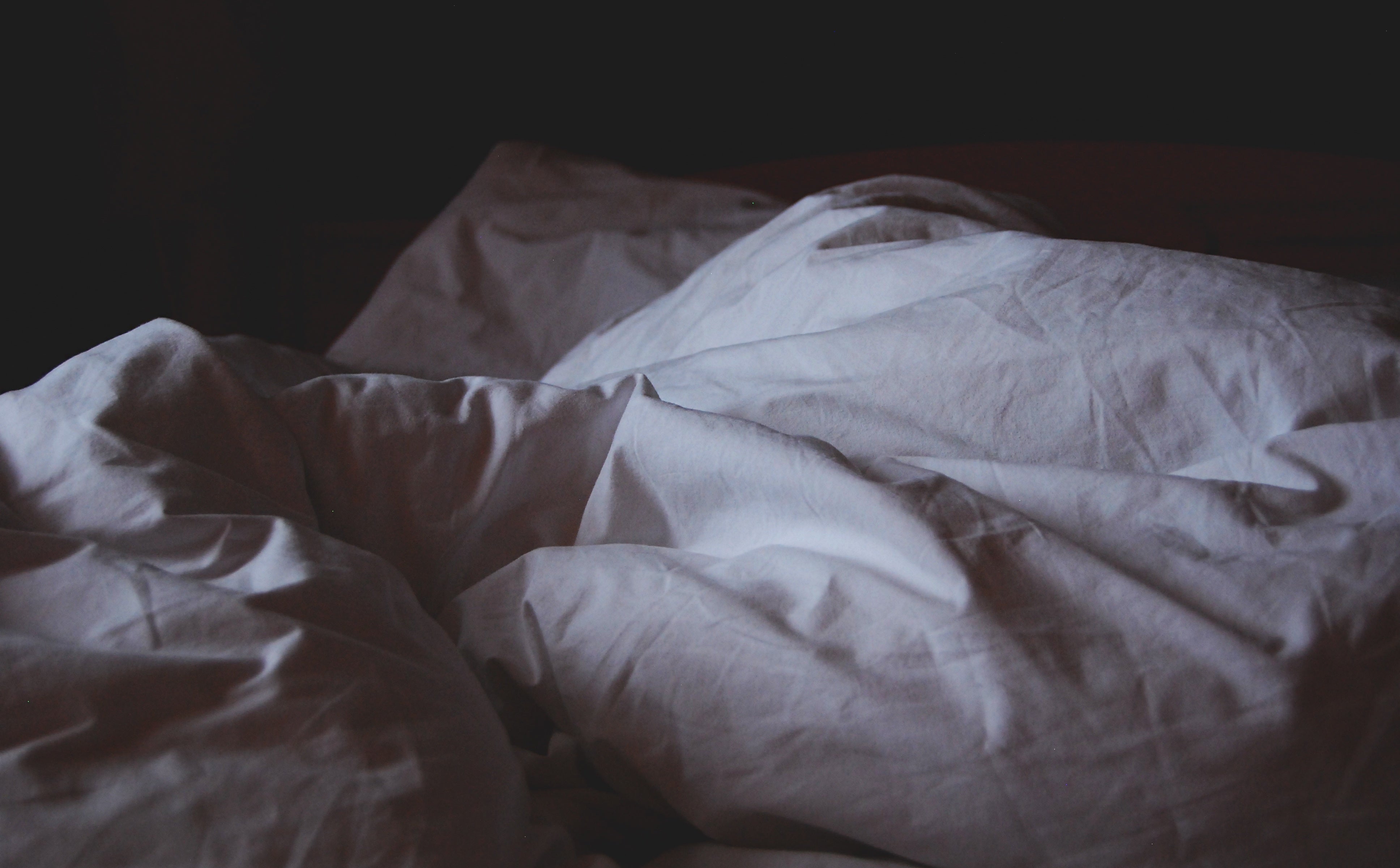 Unmade bed with rumpled white sheets in dim morning light, suggesting someone sleeping off a hangover