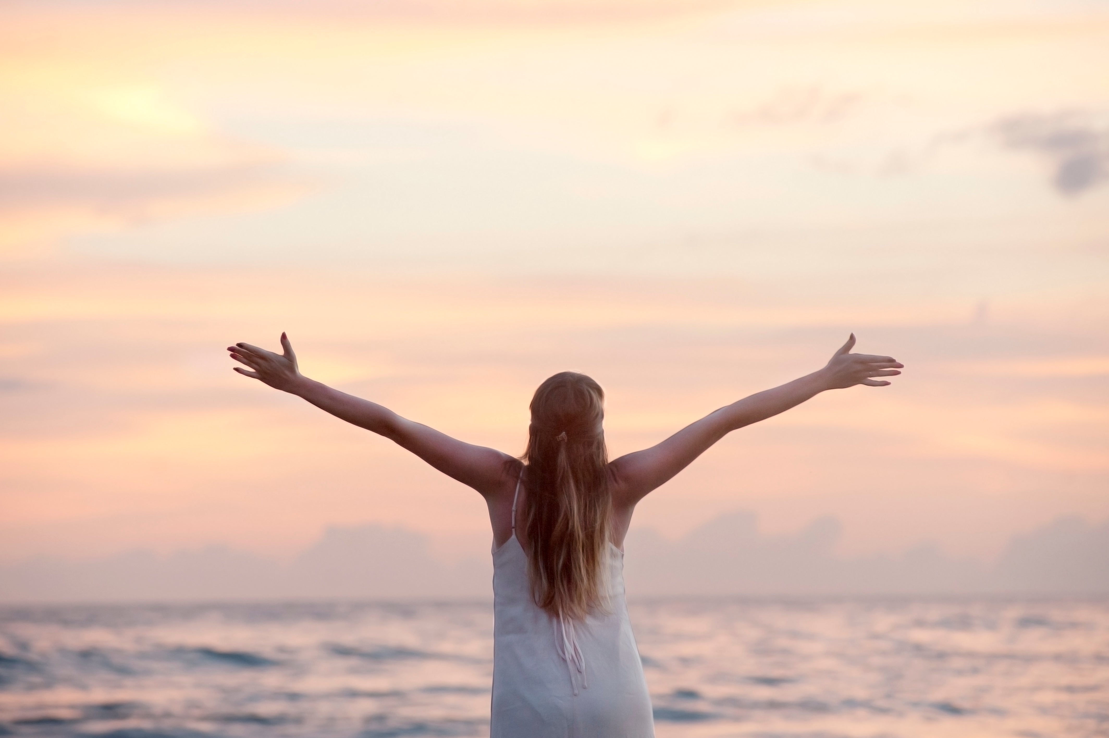 Woman standing on a beach with arms outstretched facing a pastel sunset over the ocean