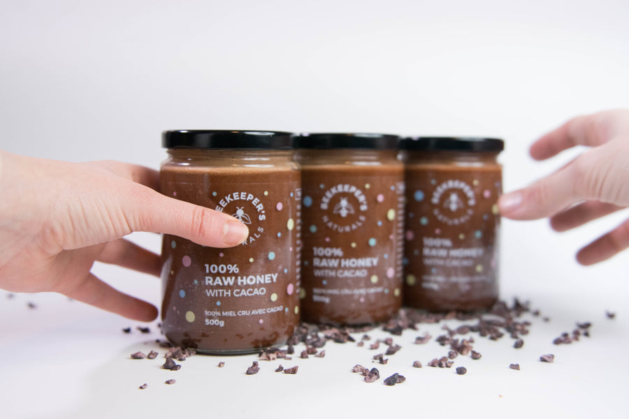 Front view of three jars of Raw Cacao Honey showcasing 100% raw honey with cacao labels and scattered cacao nibs on a white background