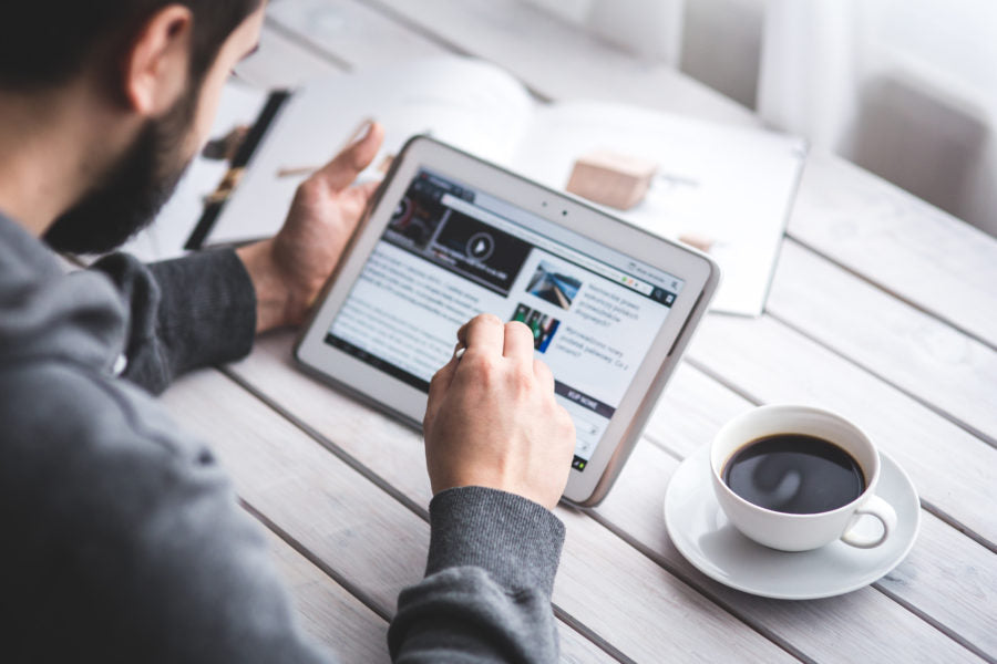Bearded man reading an article on a tablet at a white wooden table with a cup of coffee