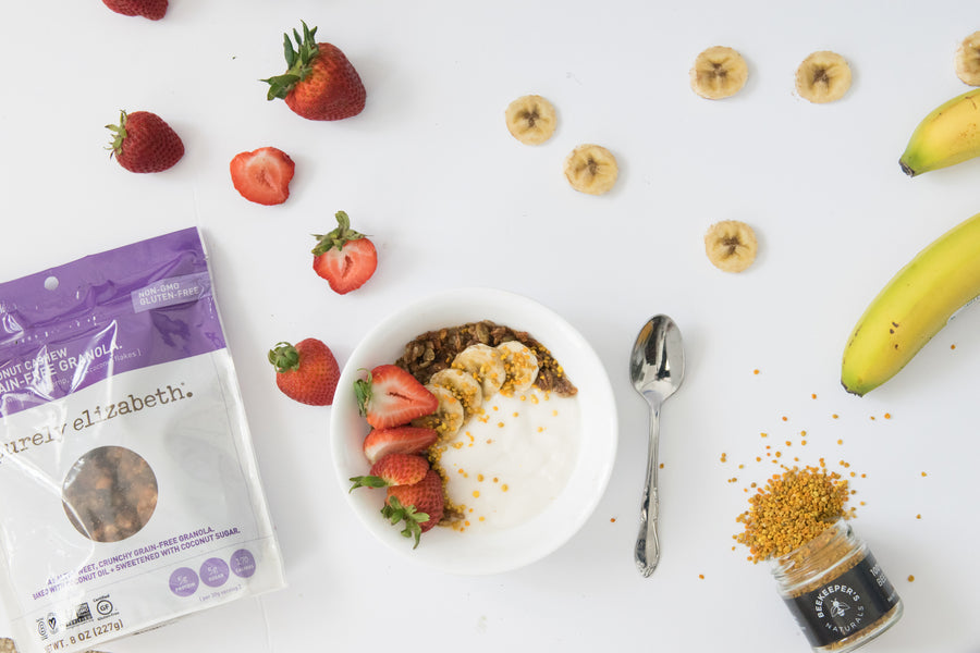 Overhead bowl of yogurt with granola, banana slices and strawberries next to a Purely Elizabeth granola pouch and jar of bee pollen