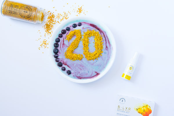 Overhead view of a purple-swirled smoothie bowl topped with bee pollen shaped as "20", a row of blueberries, a spilled pollen jar, and Beekeeper's Naturals products on a white background