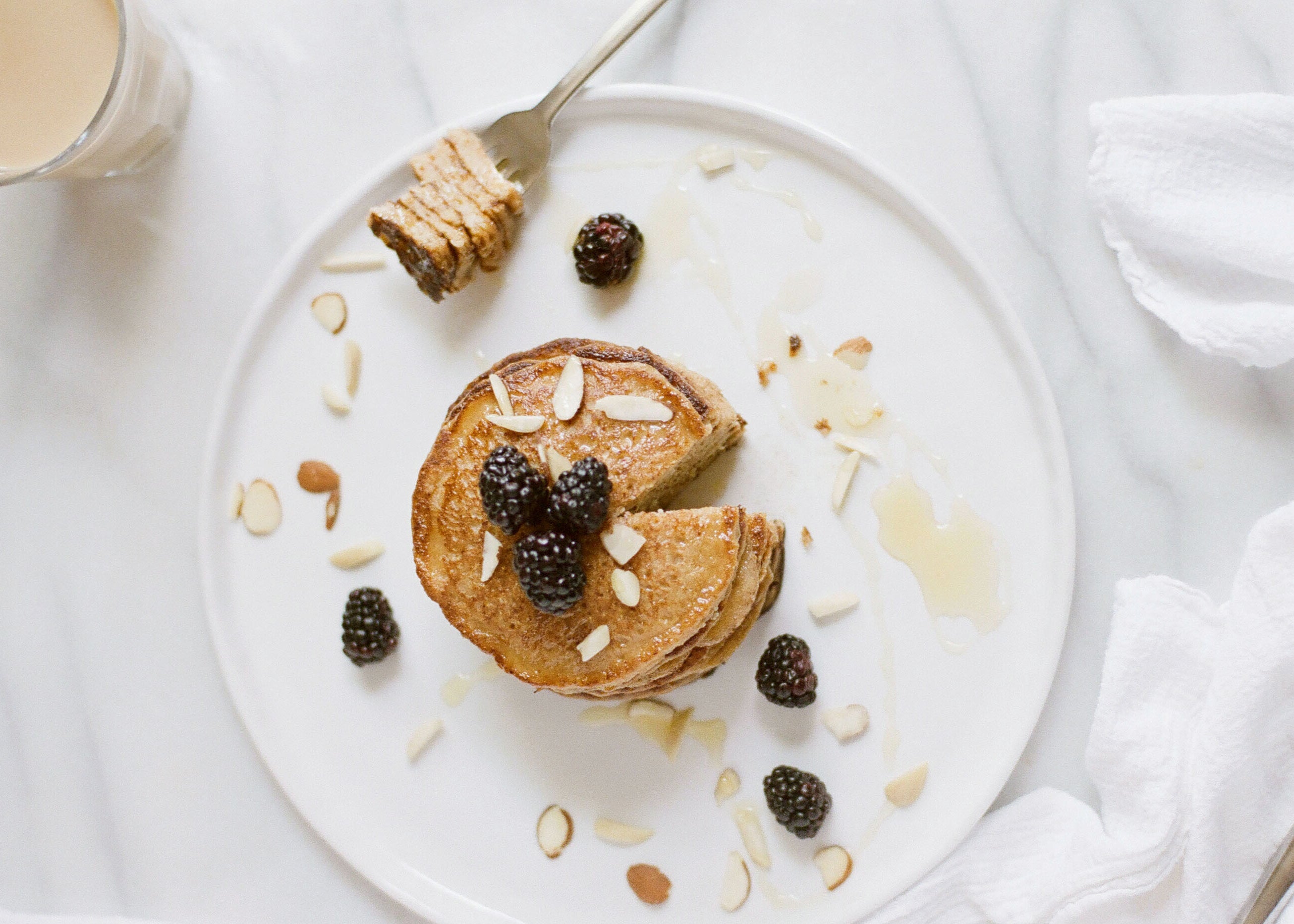Stack of banana chocolate chip pancakes topped with blackberries and sliced almonds, honey drizzle on white plate