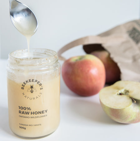 Front view of Beekeeper's Naturals 100% Raw Honey jar with honey dripping from a spoon and apples in soft-lit background