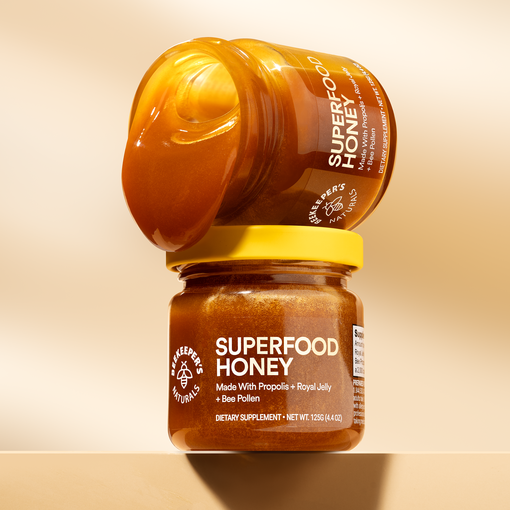 Tilted close-up of Superfood Honey jars showcasing honey pouring from the top jar and the label Superfood Honey on a warm beige background