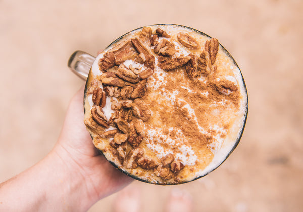 Hand holding top-down pumpkin pie smoothie bowl topped with pecans, shredded coconut and cinnamon over a blurred beige background
