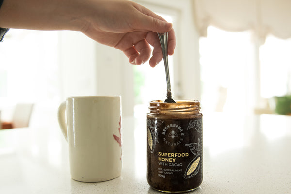 Jar of Beekeeper's Naturals Superfood Honey with Cacao being stirred with a spoon beside a coffee mug on a sunlit kitchen counter