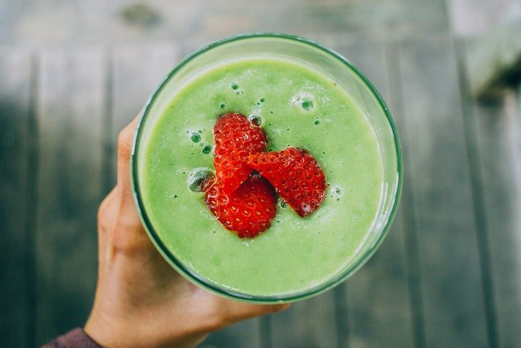 Top-down view of a hand holding a glass of green superfood smoothie topped with sliced strawberries over a wooden deck
