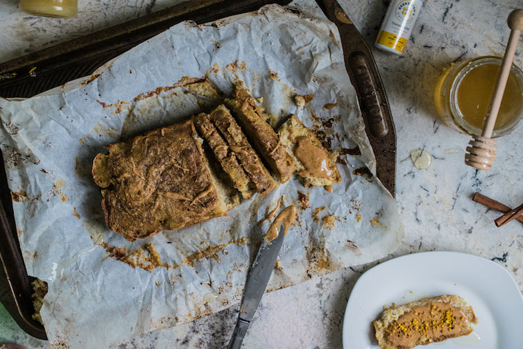 Sliced banana bread loaf on a parchment-lined baking sheet with a knife, jar of honey and a plated slice topped with nut butter on a marble countertop