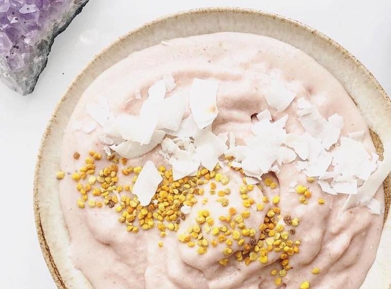 Pink smoothie bowl topped with bee pollen granules and coconut flakes on a white surface with an amethyst crystal at the rim