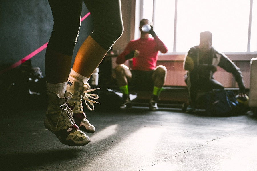 Person jumping rope in a sunlit gym with two people resting on a bench in the background