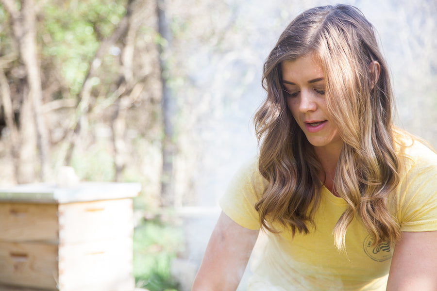 Woman tending a wooden beehive outdoors, wearing a yellow t-shirt with long wavy hair, blurred trees in background