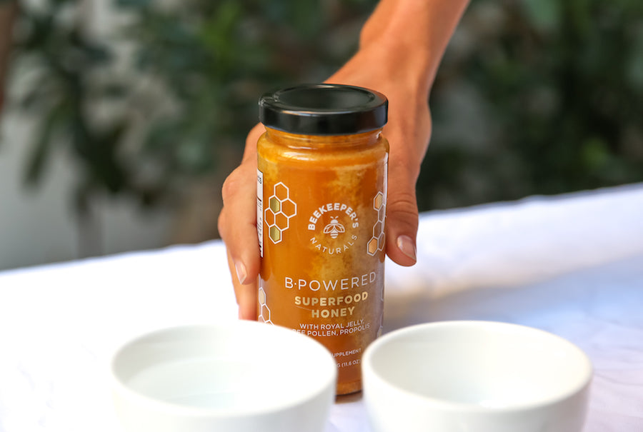 Front view of Beekeeper's Naturals B-Powered Superfood Honey jar with label visible, held by hand with white bowls in foreground and blurred plants background