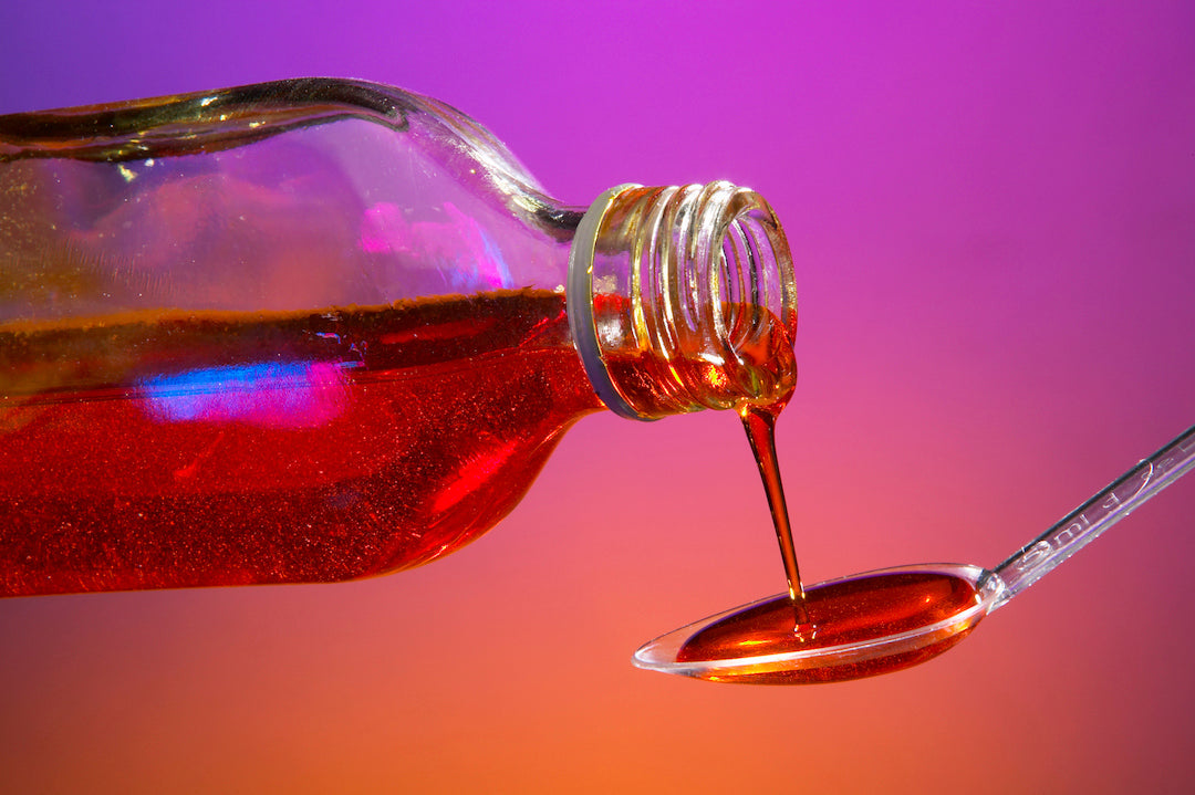 Glass medicine bottle pouring red syrup into a teaspoon against a pink-orange gradient background