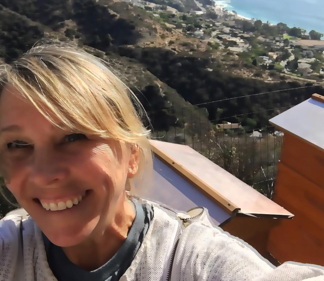 Woman smiling in a selfie beside wooden beehives on a sunny hillside overlooking the coastline