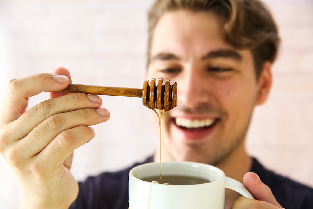 Young man smiling as he drizzles buckwheat honey from a wooden honey dipper into a white mug of tea