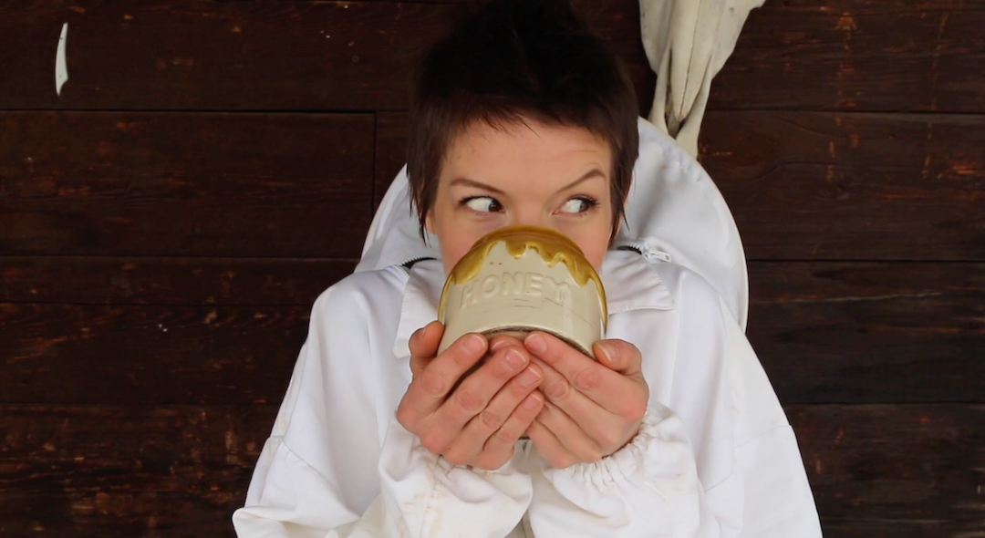 Beekeeper Laura Fayth in a white suit holding a ceramic pot labeled HONEY with honey dripping, looking to the side