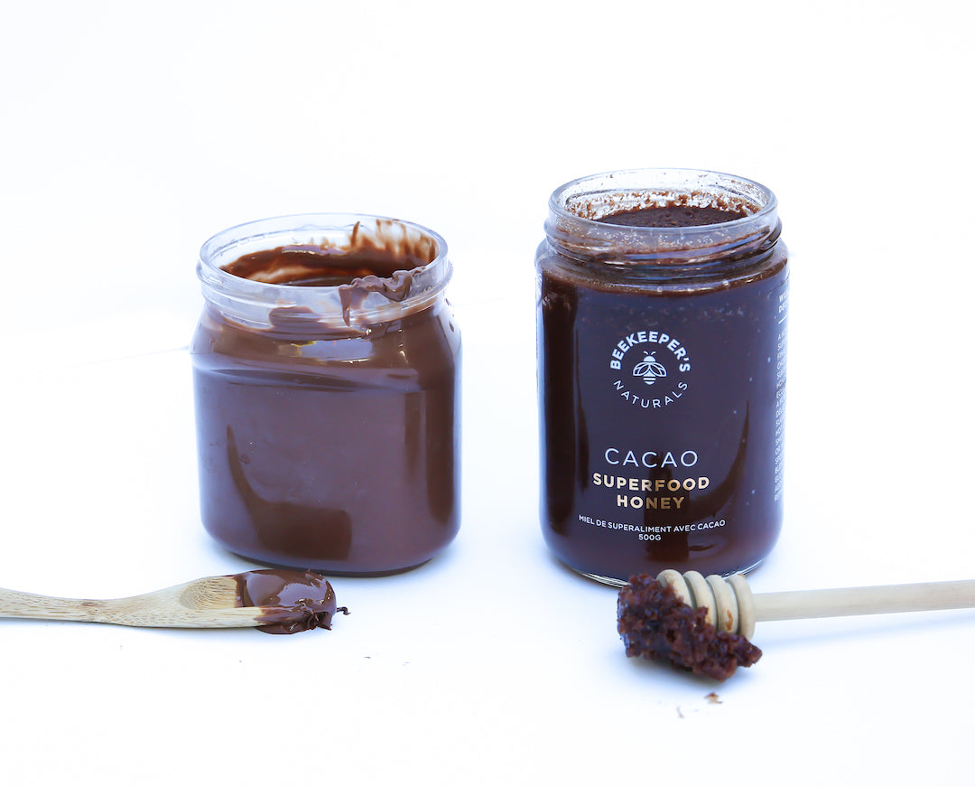 Two jars of cacao superfood honey, one open showing dark cacao-infused honey with a wooden spoon and honey dipper on white