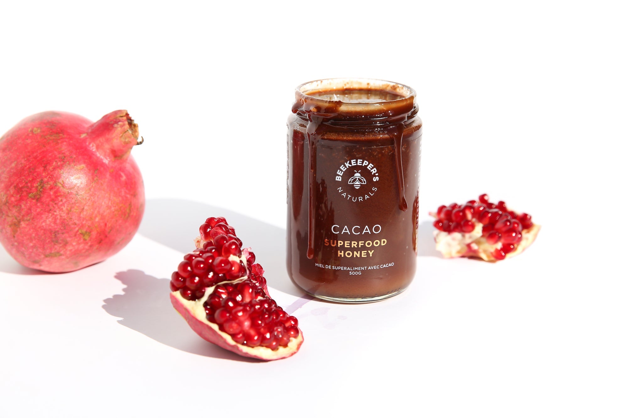 Front view of Cacao Superfood Honey jar showing cacao honey dripping from rim with pomegranate on white background