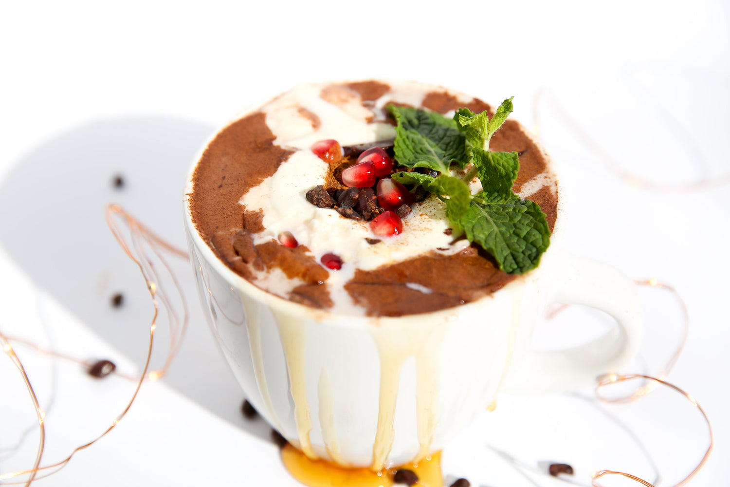 Mug of hot chocolate topped with whipped cream, pomegranate seeds, fresh mint, and honey dripping over the rim on a white background