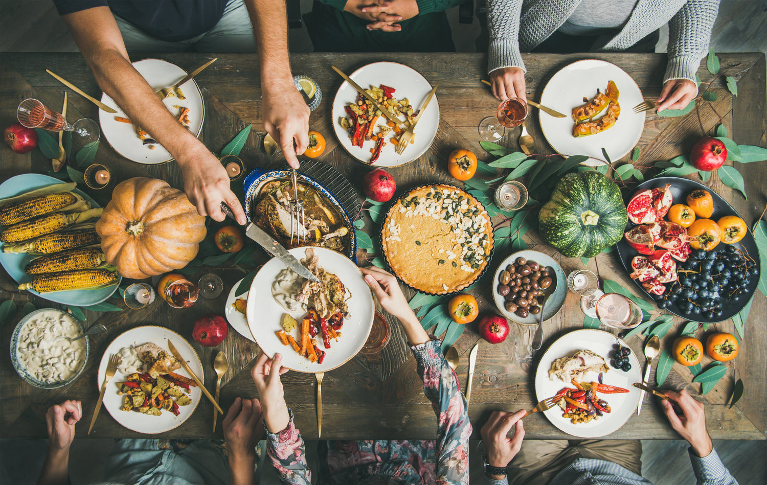 Overhead view of people serving a Thanksgiving meal with carved turkey, pumpkin pie, roasted corn, fruit, and autumn table decorations