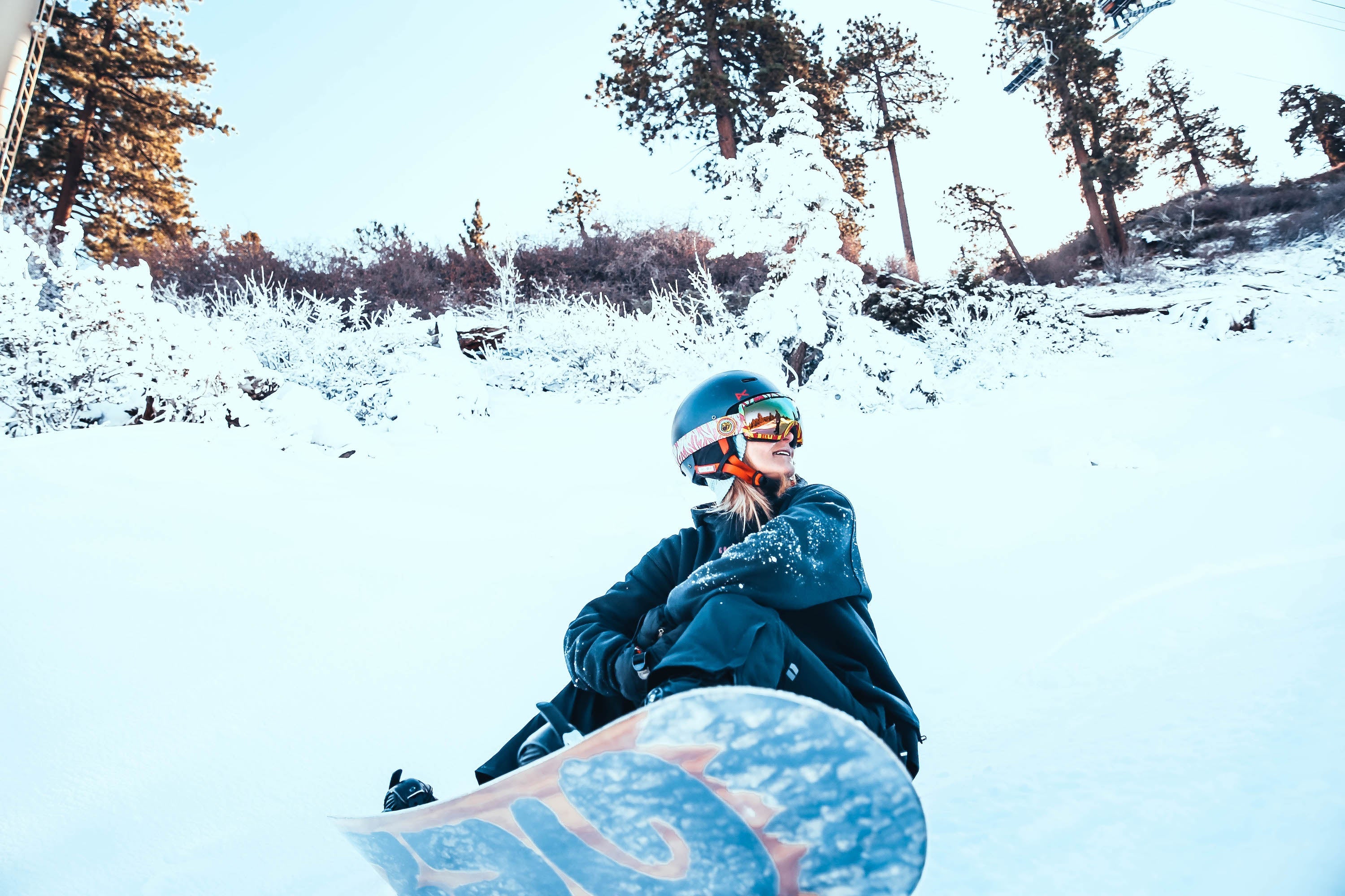 Smiling snowboarder in helmet and goggles sitting on a snowy slope with snowboard in foreground and sunlit pine trees behind