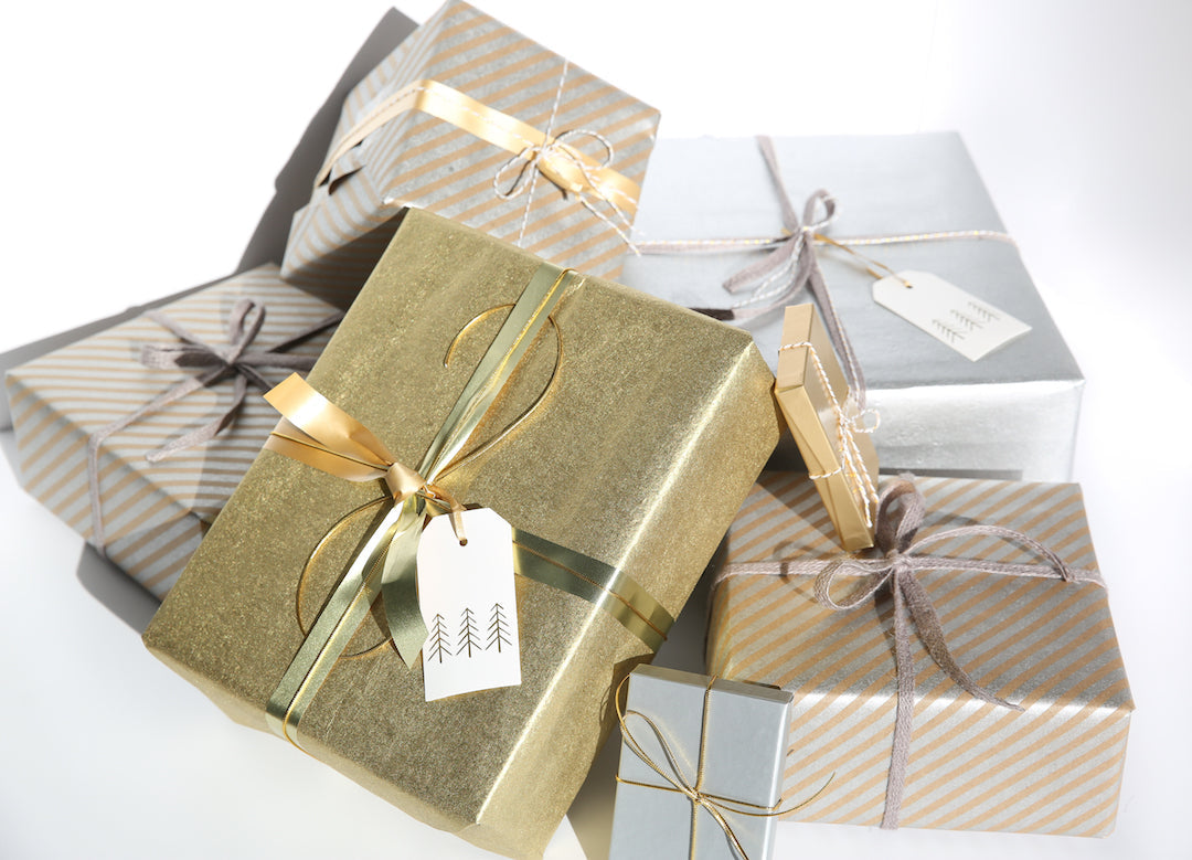 Stack of gold, silver, and striped wrapped holiday gifts with ribbons and gift tags showing small tree illustrations on a white background