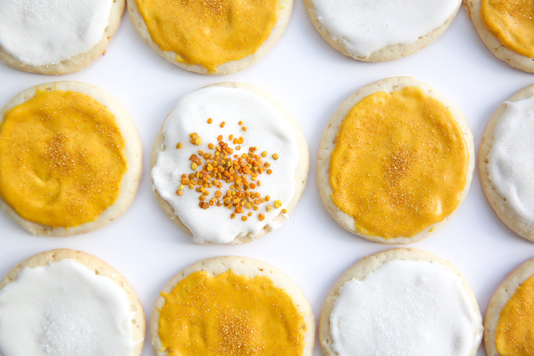 Frosted round sugar cookies arranged in rows on a white surface, alternating white and golden-yellow icing, one topped with bee pollen