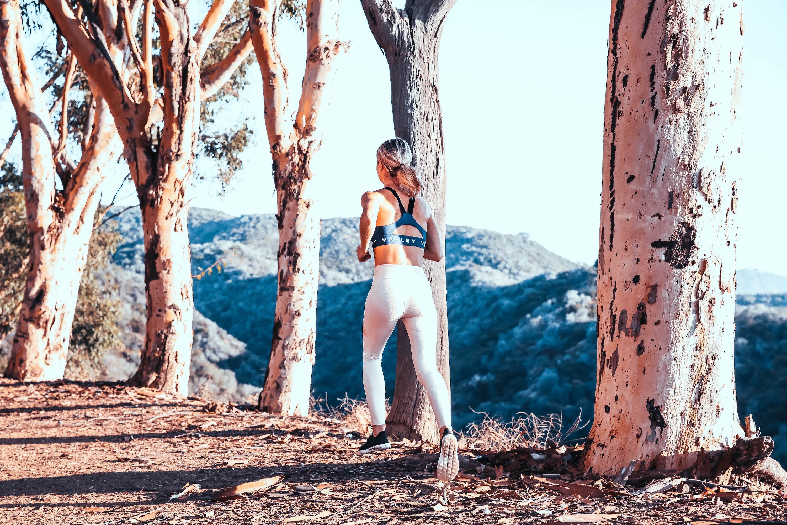 Woman jogging on a dirt trail between eucalyptus trees with mountains in the background