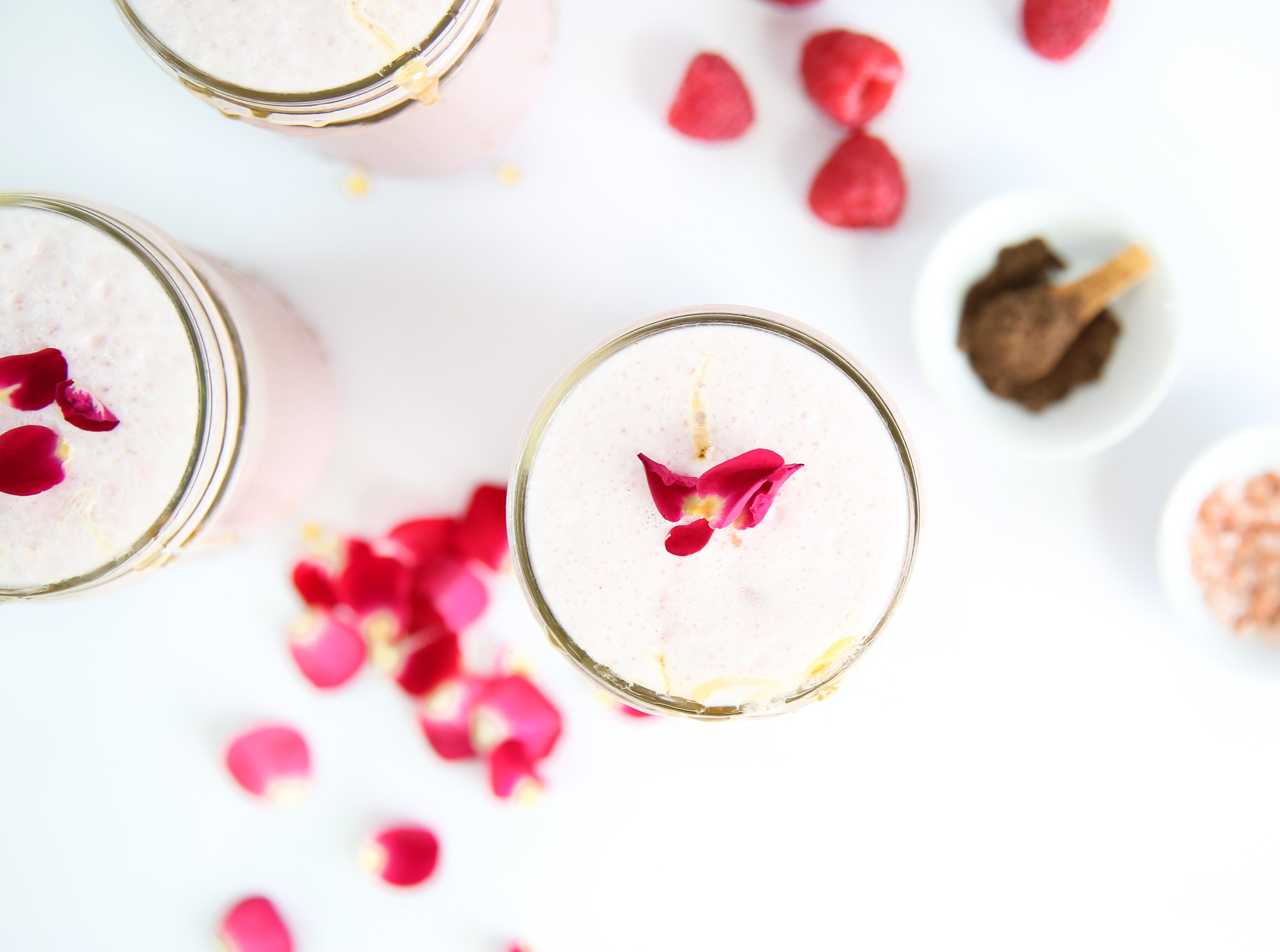 Overhead view of pink rose smoothies in glasses topped with rose petals, raspberries and spice bowls on a white background