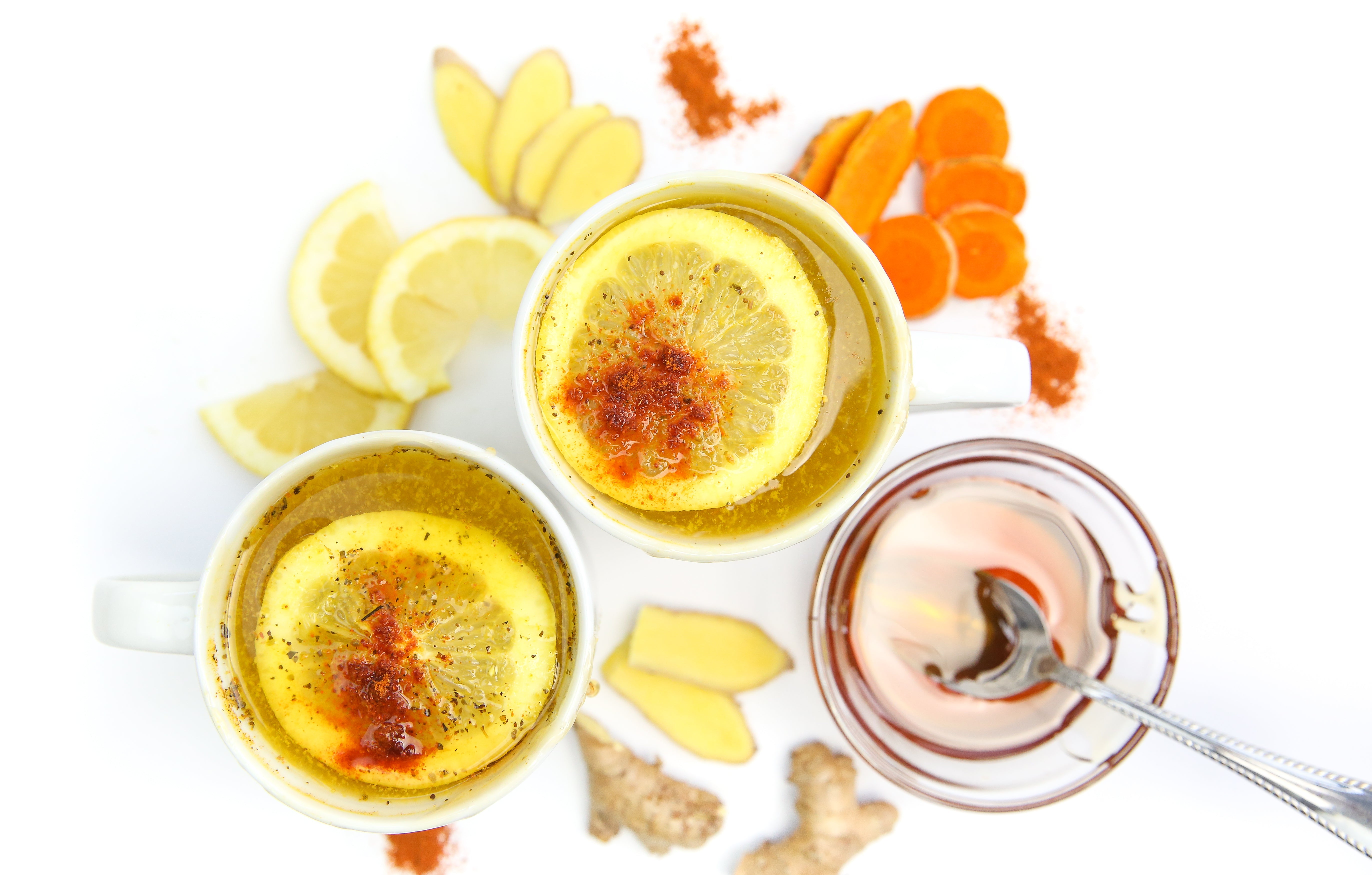 Top-down view of two hot toddy mugs with lemon slices and spices, plus honey jar and sliced ginger and turmeric on white