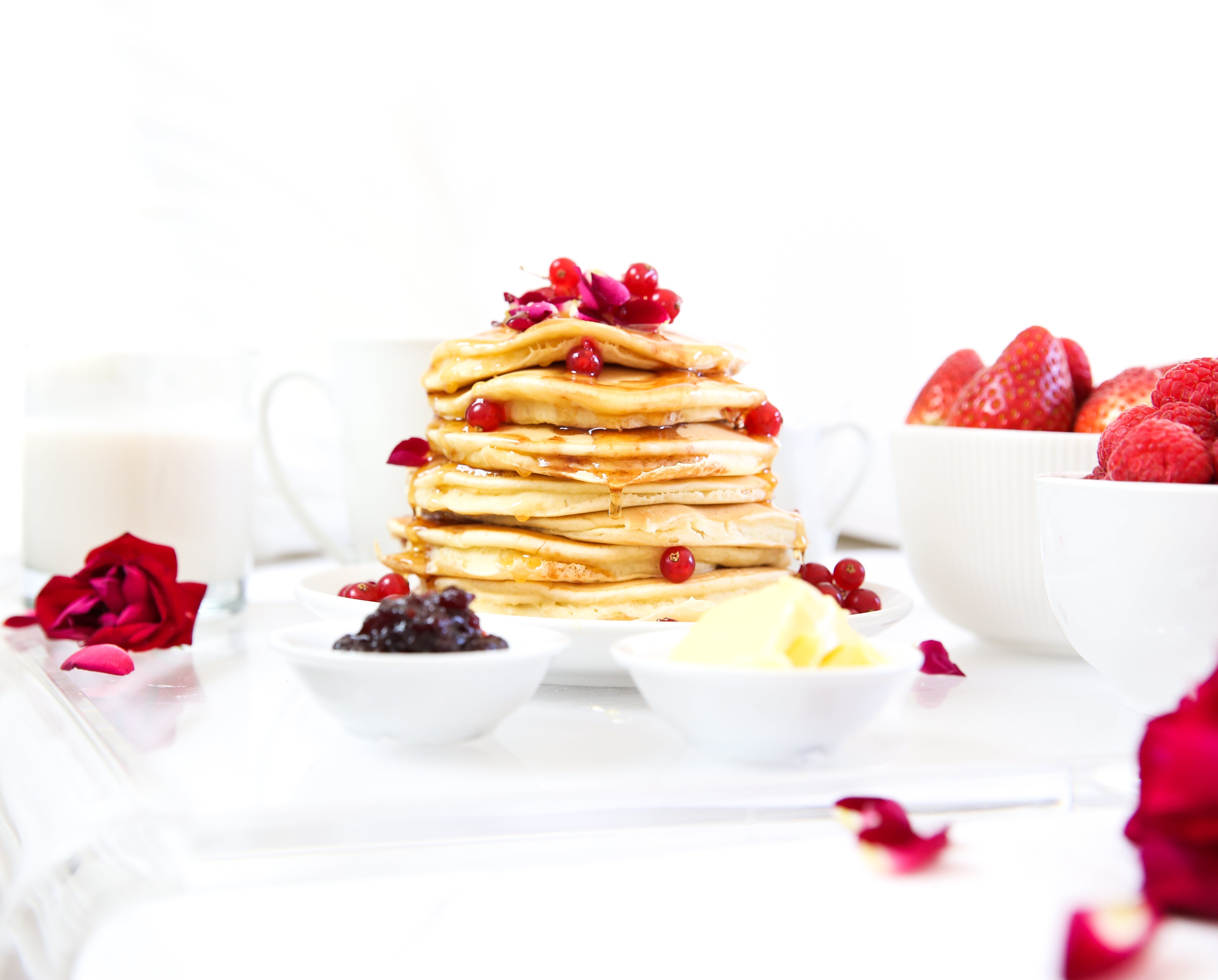 Stack of pancakes drizzled with syrup and red currants on a white tray with butter, jam, strawberries, raspberries, and rose petals
