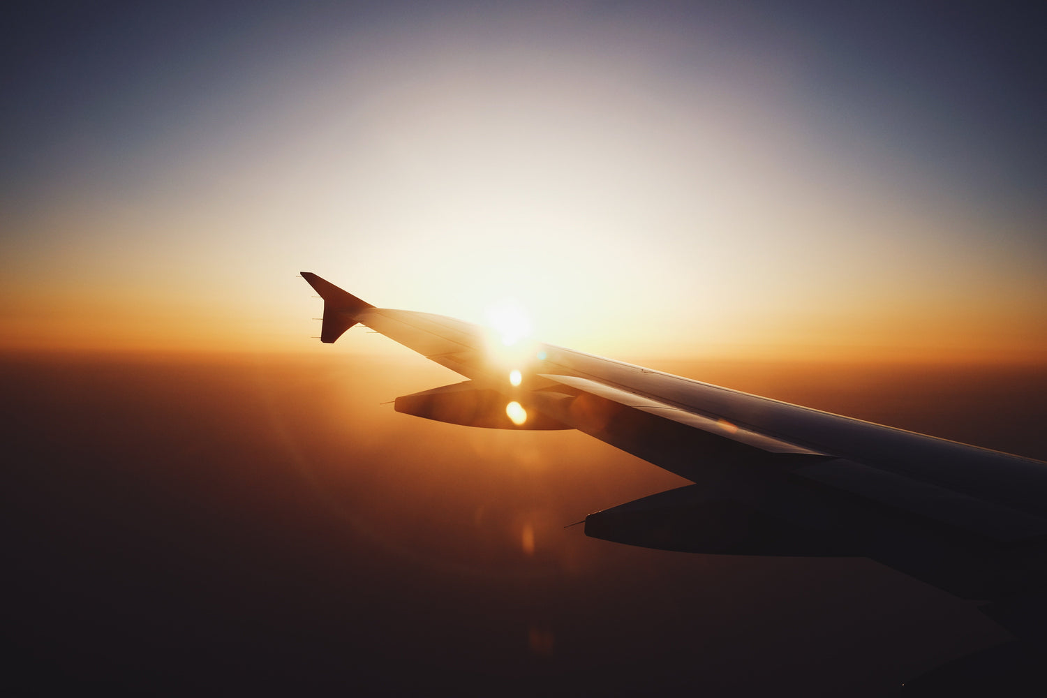 Airplane wing seen from a passenger window at sunset with the sun near the wingtip and an orange sky