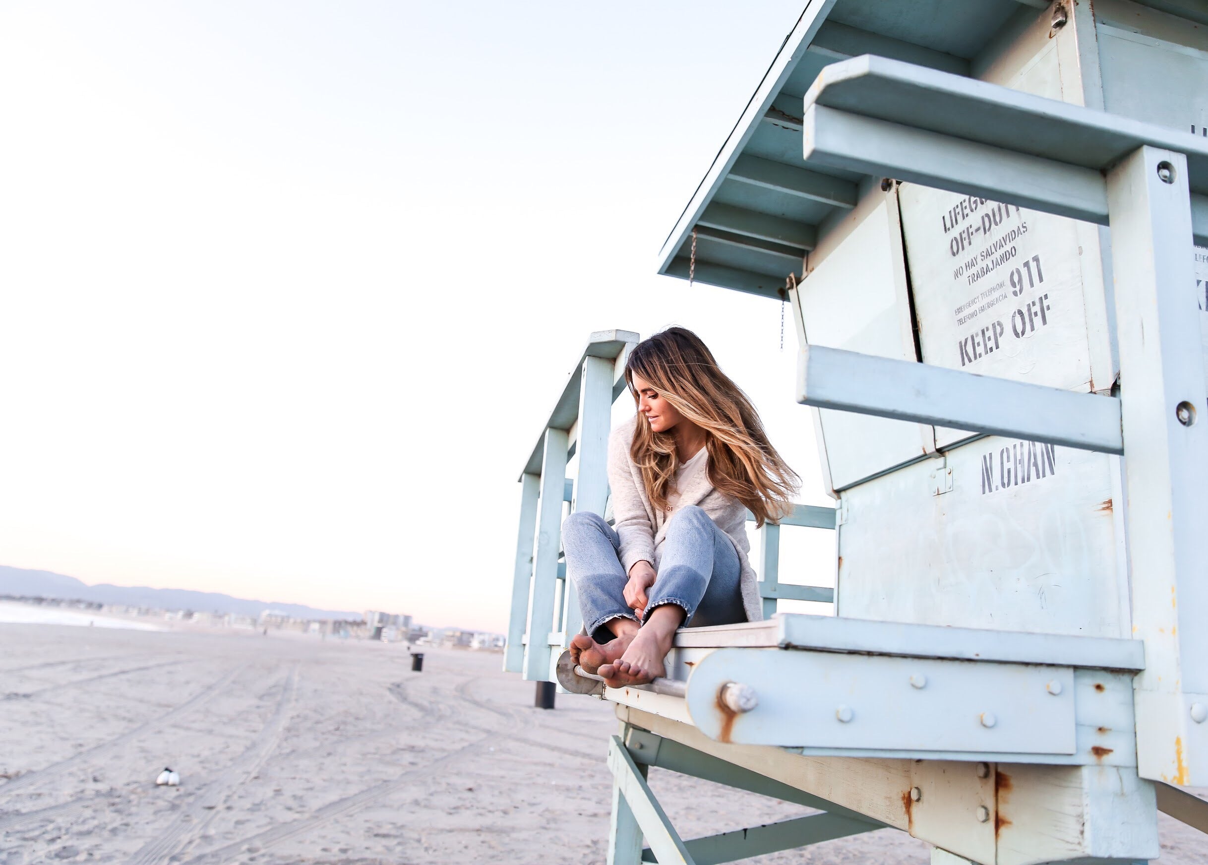 Woman sitting barefoot on a pale blue lifeguard tower overlooking an empty sandy beach at sunrise