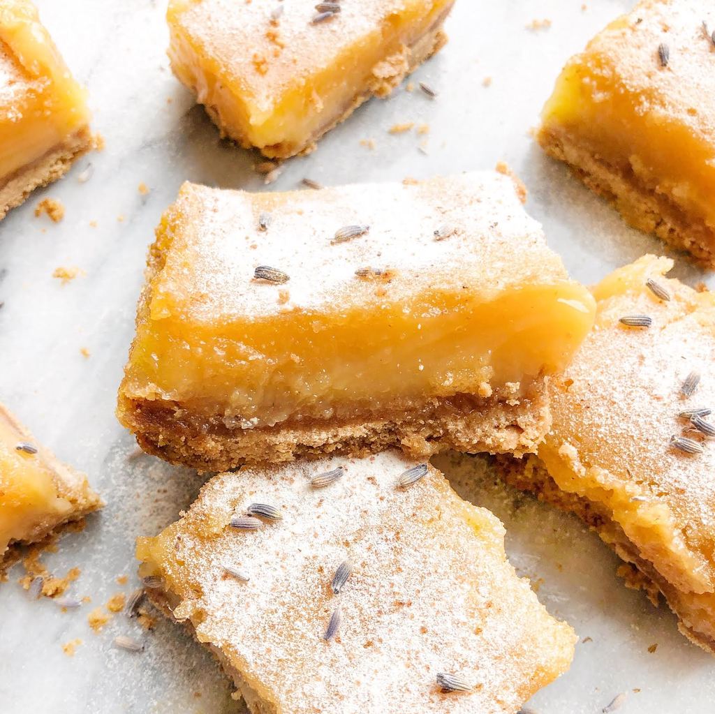 Close-up of lavender lemon bars showing glossy lemon curd layer, shortbread crust, powdered sugar and lavender buds on parchment