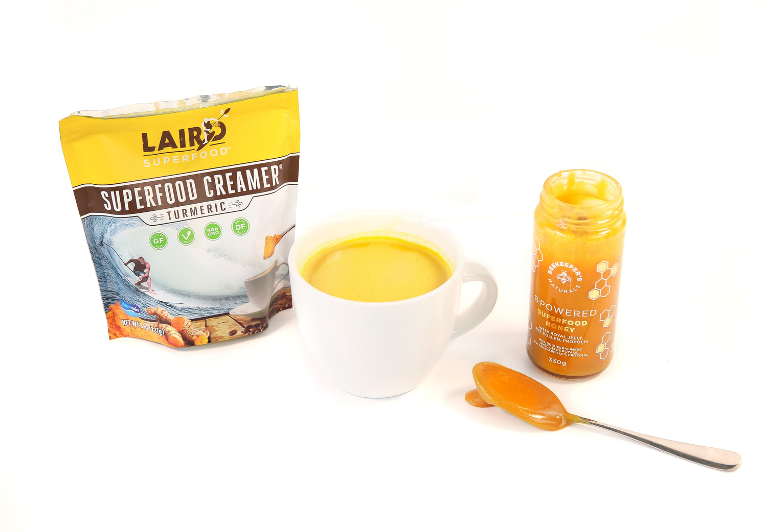 Front view of golden turmeric milk latte in a white mug with Laird Superfood turmeric creamer pouch and Beekeeper's Naturals honey jar and spoon on white background