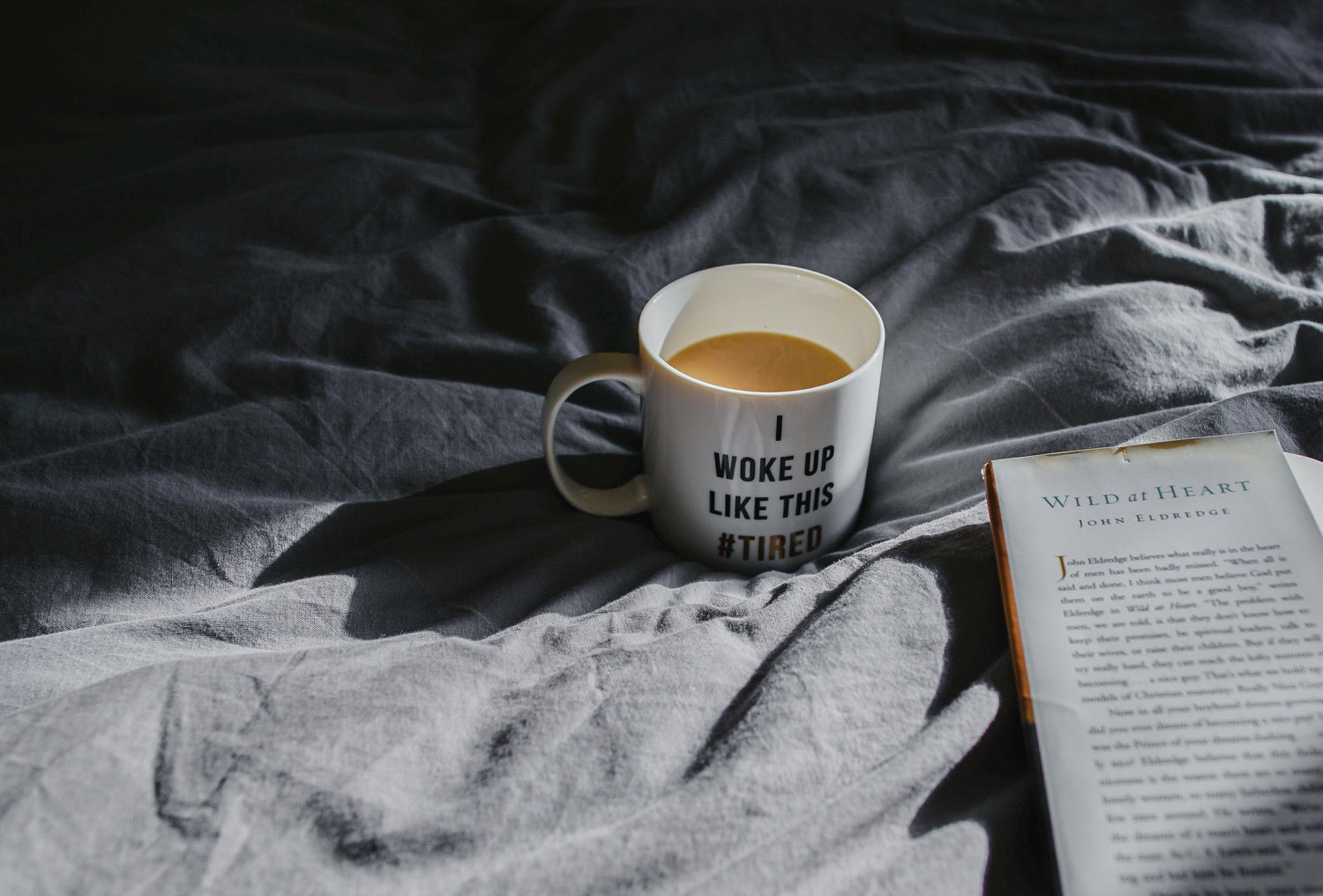 White mug of coffee reading I WOKE UP LIKE THIS #TIRED on rumpled gray sheets beside an open book in morning light